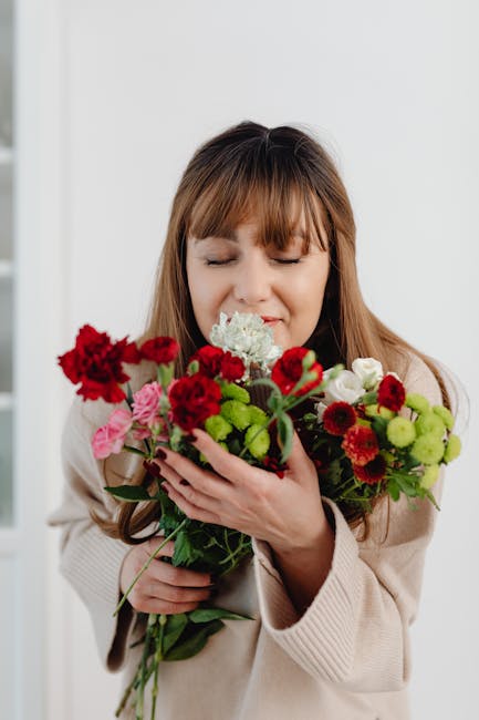 A young woman with long, light brown hair and closed eyes is gently holding a vibrant bouquet of fresh flowers close to her face, suggesting a moment of appreciation or anticipation. The bouquet featu