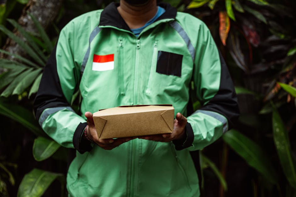 A person wearing a green and black jacket with a flag patch on the chest is holding a rectangular brown cardboard parcel, standing outdoors amidst lush green foliage and large tropical leaves. The jac