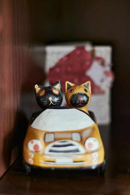 A decorative ceramic ornament featuring two black and gold cat figurines with painted facial details, sitting atop a small, orange and white toy car with a grey grille and headlights, placed on a dark