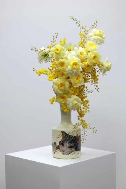 A tall, elegant floral arrangement featuring a combination of fresh, creamy yellow roses and delicate white flowers, complemented by sprigs of yellow mimosa with small, round blooms. The bouquet is ar