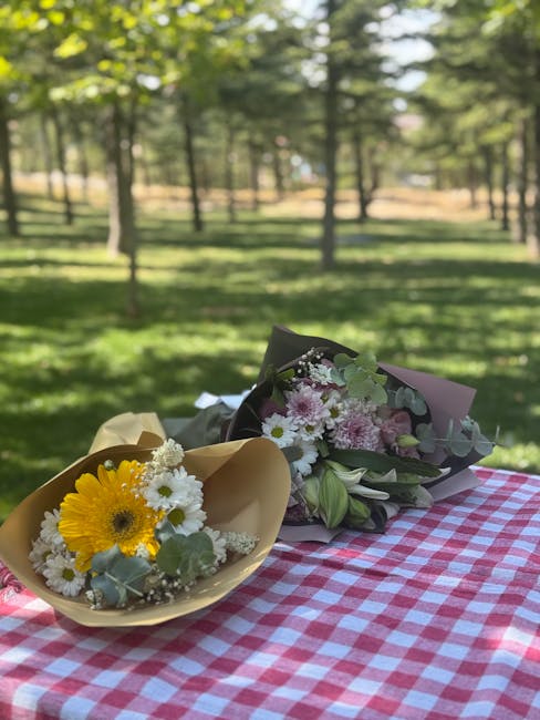 Two floral arrangements are placed on a red and white checkered tablecloth outdoors in Mountsfield Park, with lush green grass and tall trees in the background. The arrangement on the left features a 