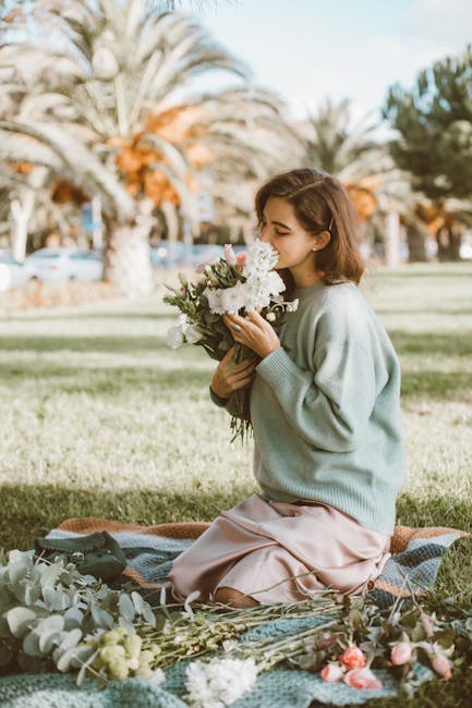 A young woman kneeling on a patterned blanket in a park with palm trees and a clear sky in the background, holding a lush bouquet of fresh flowers including white roses, hydrangeas, and greenery. She 